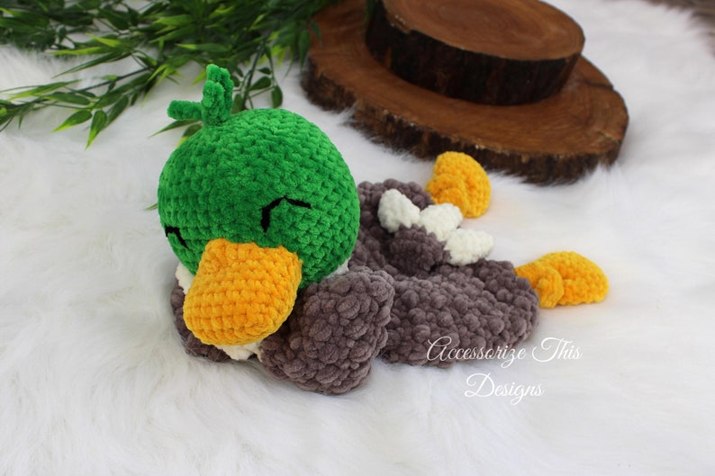 May include: A green, gray, and yellow crocheted duck plush toy with a closed eye expression. The duck is lying on its side with its head resting on its wing.