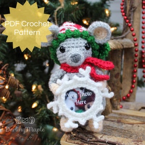 May include: A crocheted gray mouse wearing a red and white striped hat with a green wreath around its head. The mouse is holding a white crocheted snowflake with a clear photo frame that says "Your Photo Here".