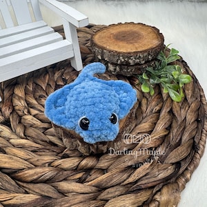 May include: A handmade, light blue crochet stingray with black safety eyes. The plush toy is displayed on a small wooden slice, set against a woven background. The stingray has a textured, fluffy appearance.