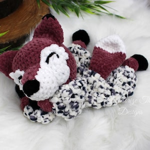 May include: A crocheted fox plush toy with a burgundy body, white face, and black eyes. The fox is lying on a white fluffy surface.