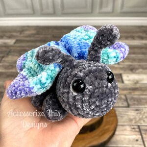 May include: A crocheted plush butterfly toy with grey fur and blue, green, and purple wings. The toy has large black eyes and a friendly expression.