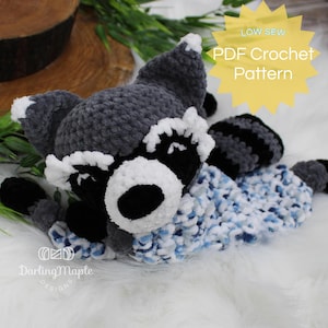 May include: Crochet pattern for a plush raccoon toy. The pattern is available as a PDF download. The raccoon is gray and black with white accents and is wearing a blue and white blanket.