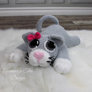 May include: A gray and white crocheted cat plush toy with pink accents. The cat has large black eyes, a pink nose, and a pink bow on its head. The cat is lying on a white fluffy surface.