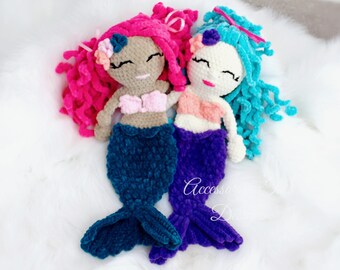 May include: Two crocheted mermaid dolls with long, curly hair. One doll has pink hair and a teal tail, the other has blue hair and a purple tail. Both dolls have a pink and white top and a brown face.