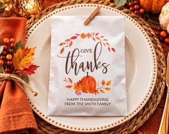 May include: A white paper favor bag featuring a printed wreath of autumn leaves, a central orange pumpkin, and the text "GIVE thanks" above "HAPPY THANKSGIVING FROM THE SMITH FAMILY". The bag is secured at the top with a small wooden clothespin.