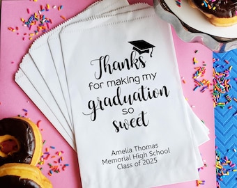May include: White paper bags with black text that reads "Thanks for making my graduation so sweet" and "Amelia Thomas Memorial High School Class of 2025". The bags are stacked on a pink surface with colorful sprinkles.