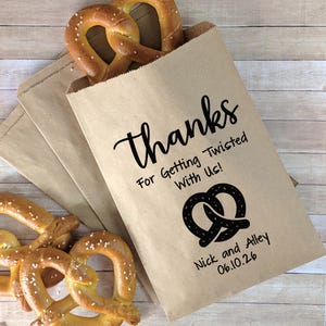 May include: Brown paper bags filled with pretzels, with the text "thanks" and "For Getting Twisted With Us!" printed in black. The bags also include a pretzel graphic and the names "Nick and Alley" with the date "06.10.26".
