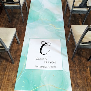 May include: A wedding aisle runner with a teal and white watercolor design and gold accents. The runner features the names "Ollie & Trayon" and the date "September 4, 2022" in a white framed section with a decorative initial.