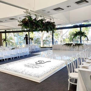 May include: A white dance floor with a custom design featuring a heart and the text "A wonderful celebration of love and laughter, Sydney & Troy, October 8, 2023". The dance floor is surrounded by white chairs and tables set for a wedding reception.