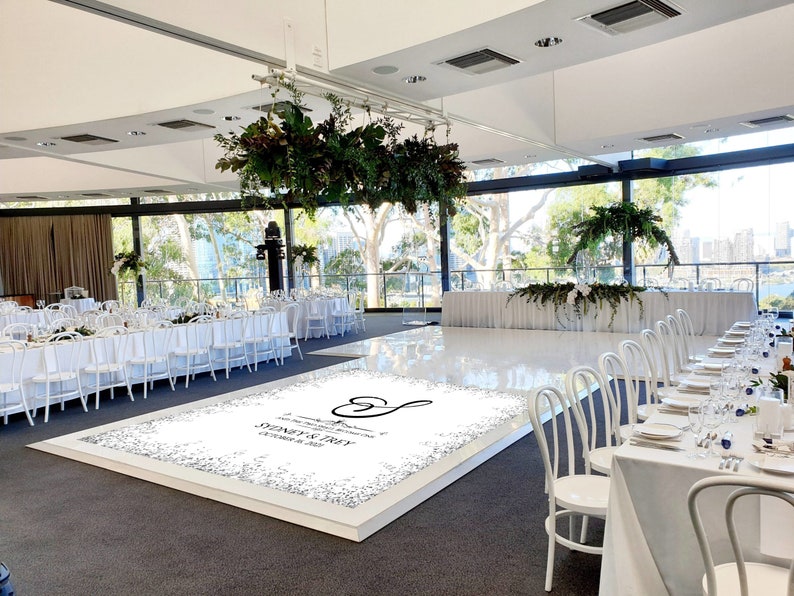 May include: A white dance floor with a custom design featuring a heart and the text "A wonderful celebration of love and laughter, Sydney & Troy, October 8, 2023". The dance floor is surrounded by white chairs and tables set for a wedding reception.