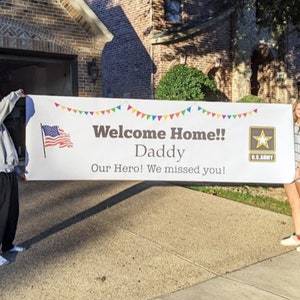 Peut inclure: Une grande banni&egrave;re blanche avec le texte "Welcome Home!! Daddy" et "Our Hero! We missed you!" est tenue par deux personnes. La banni&egrave;re comprend un drapeau am&eacute;ricain, des guirlandes color&eacute;es et un logo de l'arm&eacute;e am&eacute;ricaine.