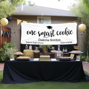 Pode incluir: Faixa de gradua&ccedil;&atilde;o branca com texto preto que diz "one smart cookie" com o nome "Dakota Gordon" e o texto "Seguin High School Class of 2025". A faixa est&aacute; pendurada acima de uma mesa preta com decora&ccedil;&otilde;es douradas e sobremesas.