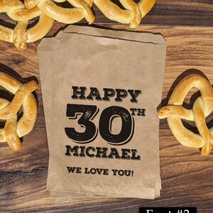 Może przedstawiać: Brązowy papierowy worek z nadrukiem "Happy 30th Michael We Love You!" w kolorze czarnym. Worek jest otoczony przekąskami w postaci precli.