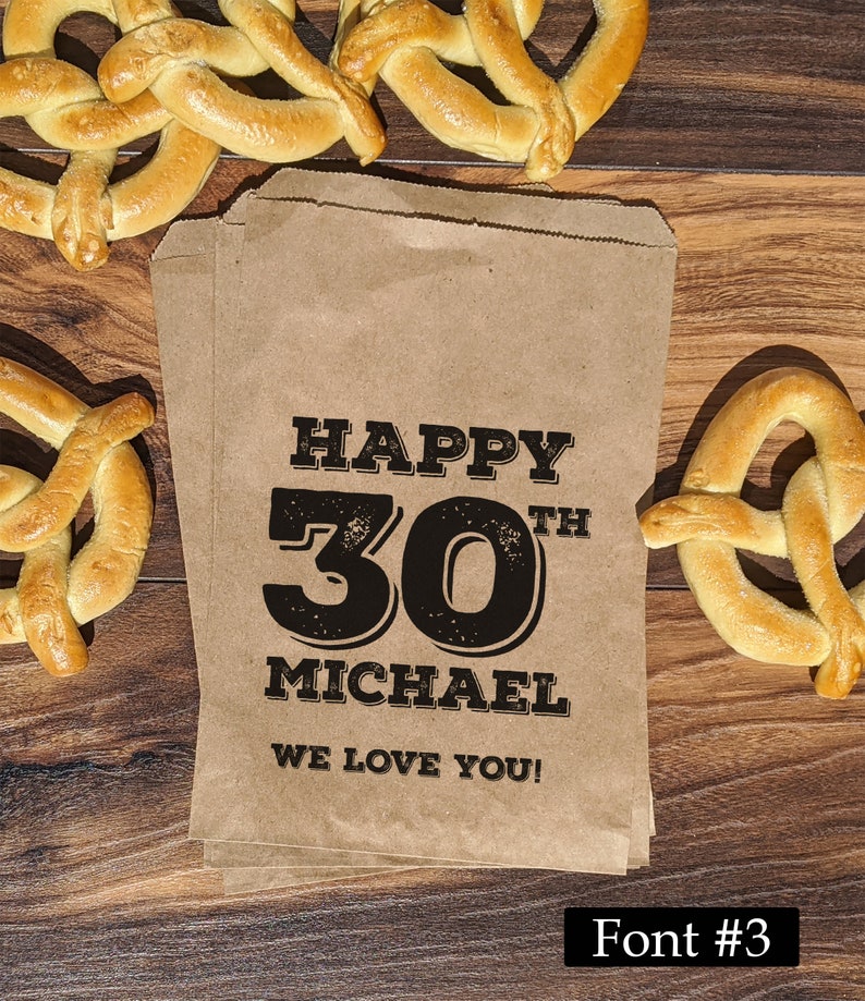 Może przedstawiać: Brązowy papierowy worek z nadrukiem "Happy 30th Michael We Love You!" w kolorze czarnym. Worek jest otoczony przekąskami w postaci precli.