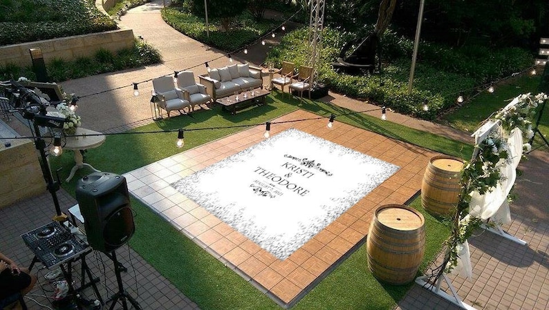 May include: A white dance floor with a custom design for a wedding. The design includes the names "Kristi & Theodore" and the date "October 2022". The dance floor is surrounded by green grass and string lights.
