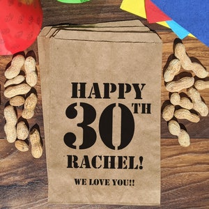Może przedstawiać: Brązowa papierowa torba z czarnym tekstem, kt&oacute;ry brzmi "Happy 30th Rachel! We love you!!" Torba jest otoczona orzeszkami ziemnymi i innymi dekoracjami imprezowymi.