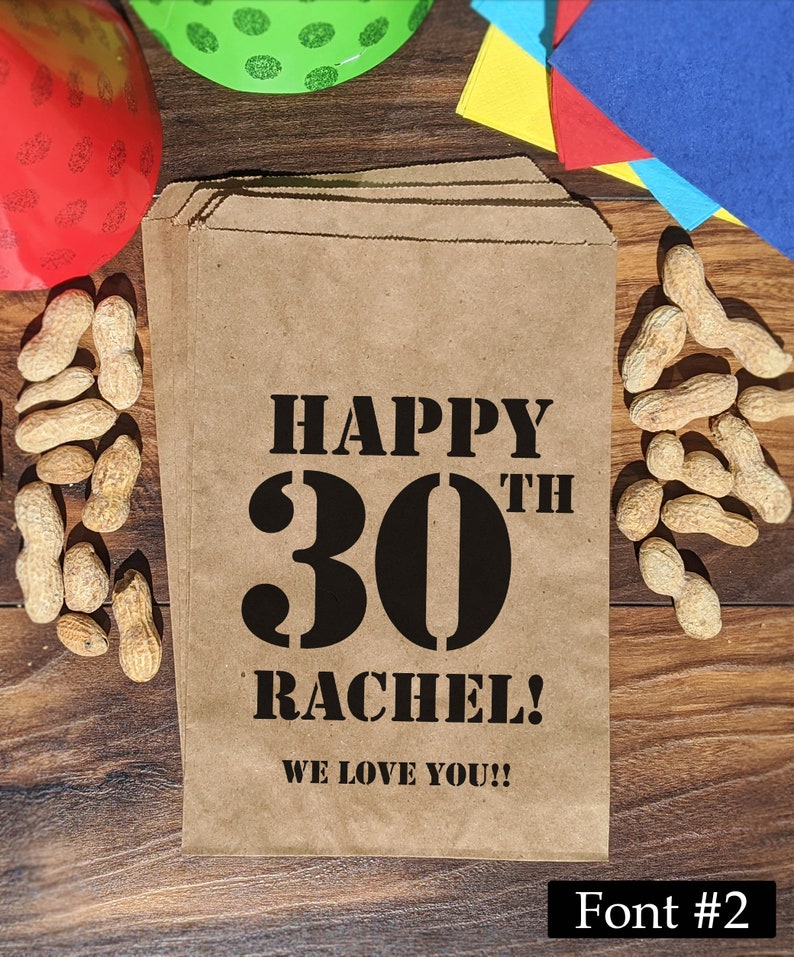 Może przedstawiać: Brązowa papierowa torba z czarnym tekstem, kt&oacute;ry brzmi "Happy 30th Rachel! We love you!!" Torba jest otoczona orzeszkami ziemnymi i innymi dekoracjami imprezowymi.