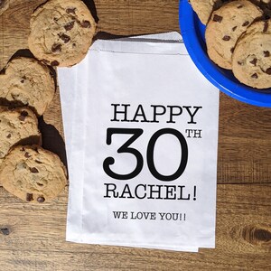 Może przedstawiać: Biały papierowy worek z czarnym tekstem, kt&oacute;ry brzmi "Happy 30th Rachel! We love you!!" Worek leży na drewnianej powierzchni z ciasteczkami z kawałkami czekolady.
