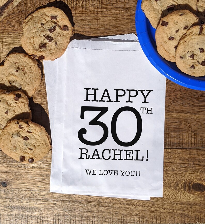 Może przedstawiać: Biały papierowy worek z czarnym tekstem, kt&oacute;ry brzmi "Happy 30th Rachel! We love you!!" Worek leży na drewnianej powierzchni z ciasteczkami z kawałkami czekolady.