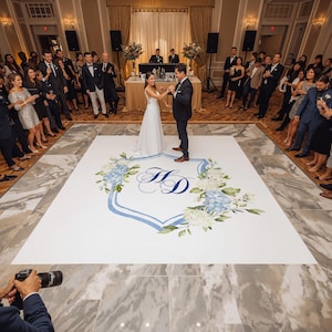 May include: A large square white dance floor decal featuring a blue crest outline, blue and white floral accents, and the monogram letters HD in the center. The decal is applied to a marble-patterned floor in a ballroom setting with guests.