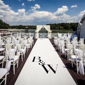 May include: White wedding ceremony chairs are set up on a wooden dock overlooking a lake. A white aisle runner with the initials "B&W" leads to a white wedding arch decorated with white flowers.