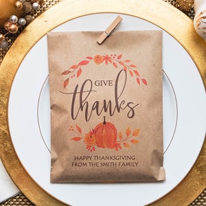 May include: A brown paper bag with the words "GIVE thanks" and a pumpkin illustration, clipped to a white plate with a gold rim. The bag also includes the text "HAPPY THANKSGIVING FROM THE SMITH FAMILY".