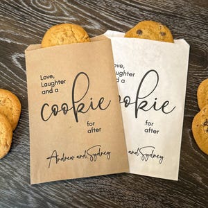 Op de afbeelding: Twee papieren koekjeszakjes, &eacute;&eacute;n bruin en &eacute;&eacute;n wit, elk met een koekje erin. De zakjes hebben de tekst "Love, Laughter and a Cookie for after" en de namen "Andrew and Sydney" erop gedrukt. Er liggen meerdere koekjes.