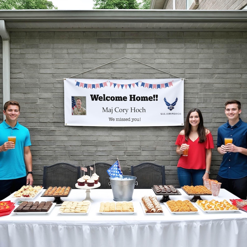 Peut inclure: Une banni&egrave;re avec l'inscription "Welcome Home!!" avec le nom "Maj Cory Hoch" et le logo de l'US Air Force. Une table est dress&eacute;e avec divers desserts et boissons, avec trois personnes devant la table.