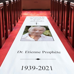 May include: A white aisle runner with a black border and text that reads "In loving the life of living, of our dear pastor, Dr. Etienne Prophete 1939-2021". A photo of a man is in the center of the runner.