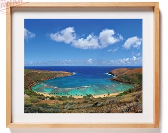 May include: A framed landscape photograph of Hanauma Bay in Hawaii, featuring a crescent-shaped beach with turquoise water, surrounded by volcanic hills under a bright blue sky with white clouds. The wooden frame has a light natural finish.
