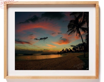 May include: A framed landscape photograph of a beach at sunset with a vibrant orange and blue sky, dark silhouettes of palm trees, and calm ocean waters. The print is encased in a light-colored wooden frame with a white mat border.