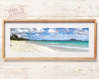 May include: A panoramic framed photograph of a tropical beach with white sand, turquoise ocean water, and a blue sky filled with white clouds. The wooden frame surrounds a white mat. The text MADE IN HAWAII is printed in the upper left corner.