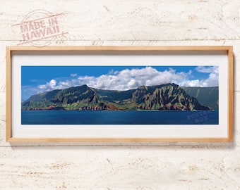 May include: A panoramic landscape photograph in a light wood frame featuring the green, jagged cliffs of the Na Pali Coast in Hawaii rising above deep blue ocean waters under a bright blue sky with white clouds. The text MADE IN HAWAII is visible.