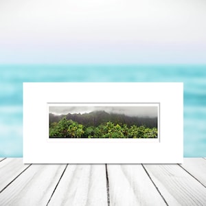 May include: A framed photograph of a lush green mountain range with clouds in the distance. The photograph is set against a blurred background of a turquoise ocean.