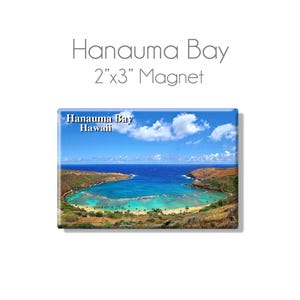 May include: A 2x3 inch magnet featuring a scenic view of Hanauma Bay, Hawaii. The image shows a crescent-shaped bay with turquoise water, surrounded by lush green hills and a bright blue sky with fluffy white clouds.