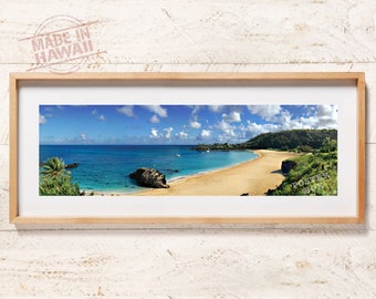 May include: A panoramic landscape photograph in a light wood frame featuring a sandy beach, turquoise ocean water, a large dark rock formation, and green hills under a bright blue sky with white clouds. The text MADE IN HAWAII is visible.