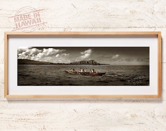 May include: A panoramic photograph of a group of people paddling a canoe in the ocean with a mountain in the background. The sky is cloudy and the water is calm. The image is in sepia tone.