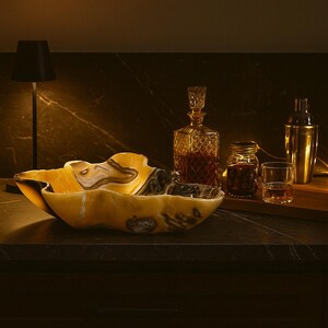May include: A decorative onyx bowl with a unique, organic shape, featuring shades of yellow, brown, and black. A crystal decanter, a cocktail shaker, and a small glass are also visible, suggesting a bar or home decor theme.