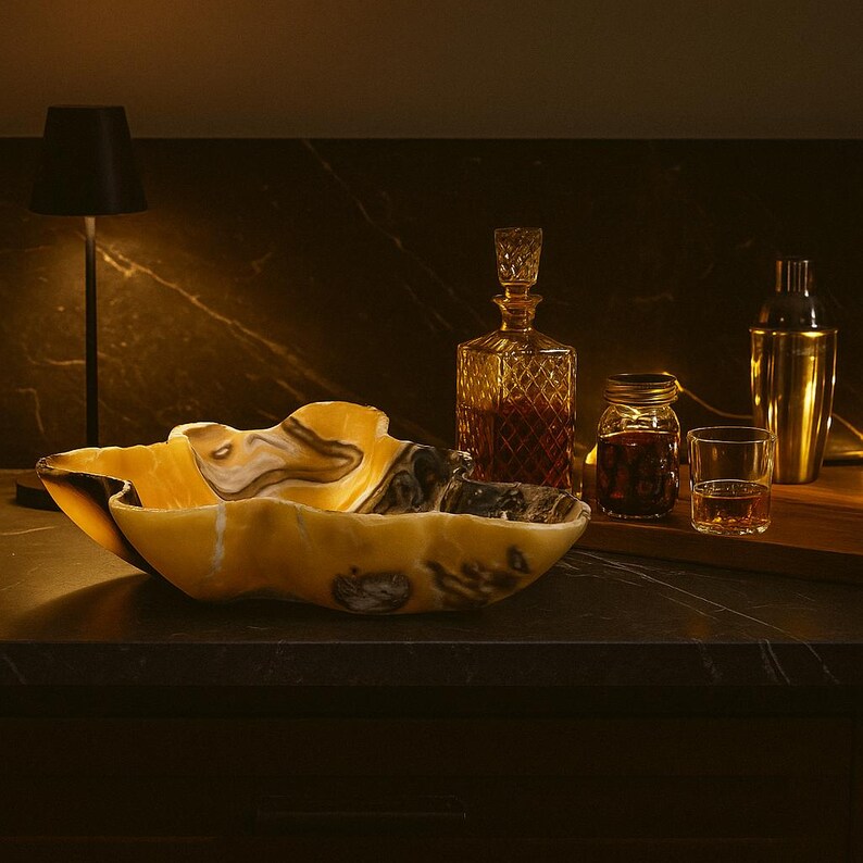 May include: A decorative onyx bowl with a unique, organic shape, featuring shades of yellow, brown, and black. A crystal decanter, a cocktail shaker, and a small glass are also visible, suggesting a bar or home decor theme.