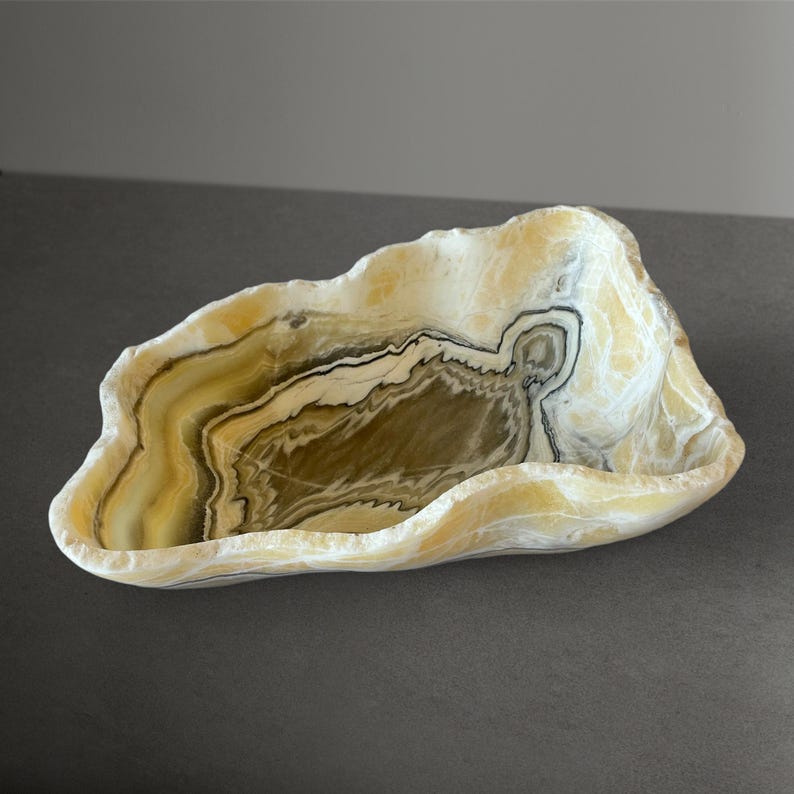 May include: A shallow, irregular bowl carved from natural stone featuring translucent layers of cream, golden yellow, and dark brown banding. The edges are rough and organic, showcasing the raw, polished texture of the mineral formation.
