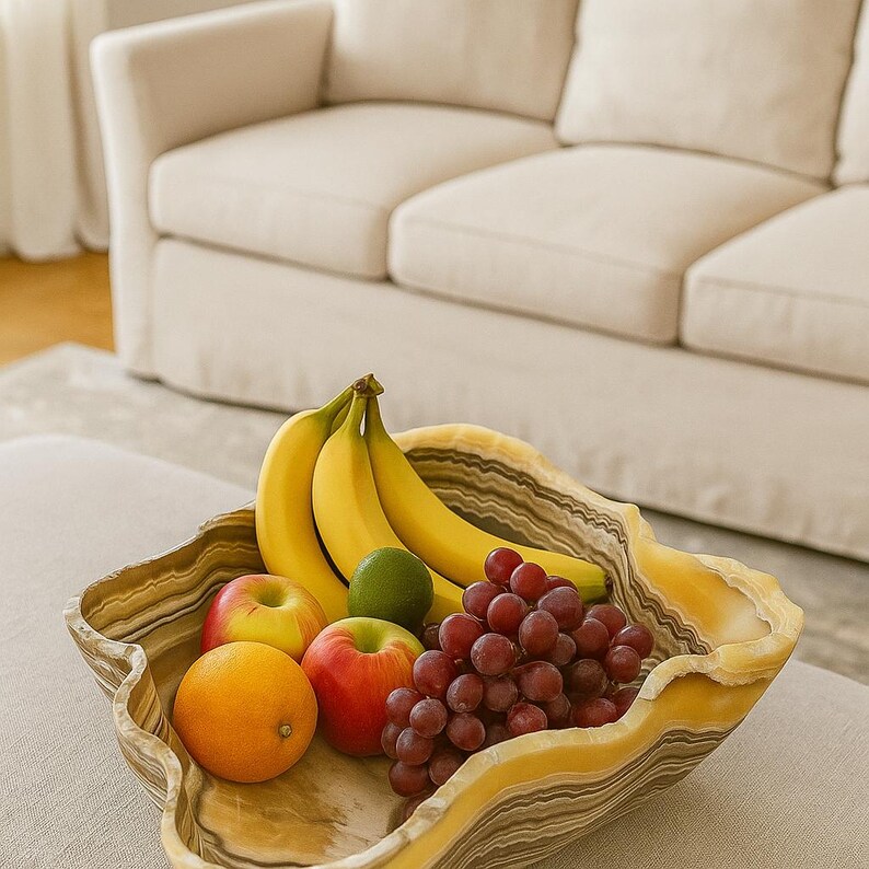 May include: A decorative fruit bowl filled with fresh fruit. The bowl has a unique, organic shape with yellow and brown tones. It holds bananas, red grapes, apples, an orange, and a lime, creating a colorful display.