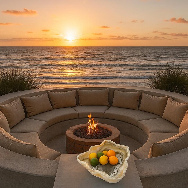 May include: A circular outdoor seating area with beige cushions and a central fire pit. The scene is set against a backdrop of the ocean at sunset. A decorative bowl with citrus fruits sits on a table in the foreground.