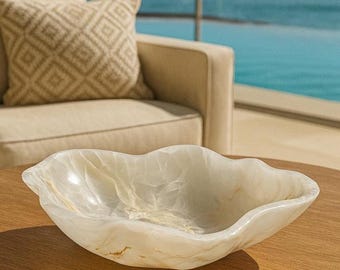 May include: A decorative, off-white onyx bowl with a wavy rim sits on a wooden table. The bowl has a translucent quality with subtle veining. A beige armchair and patterned pillow are in the background, with a view of the ocean.