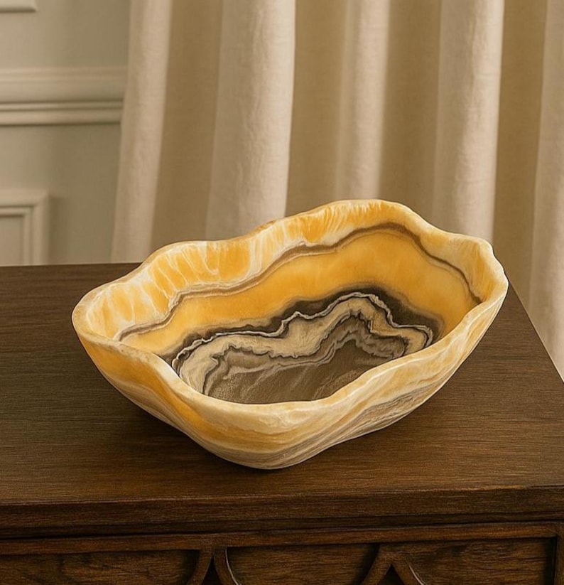 May include: A decorative bowl crafted from natural stone, featuring organic, wavy edges and a polished finish. The bowl displays a striking color palette of translucent golden yellow, cream, and deep charcoal gray, arranged in swirling, layered bands.