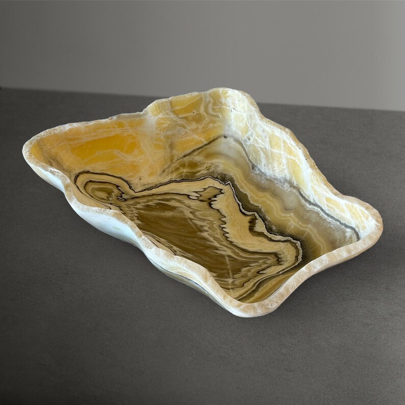 May include: A decorative onyx bowl with a unique, organic shape. The bowl features a blend of yellow, brown, and white tones, with intricate veining patterns throughout. The edges have a natural, uneven contour, adding to its handcrafted appearance.