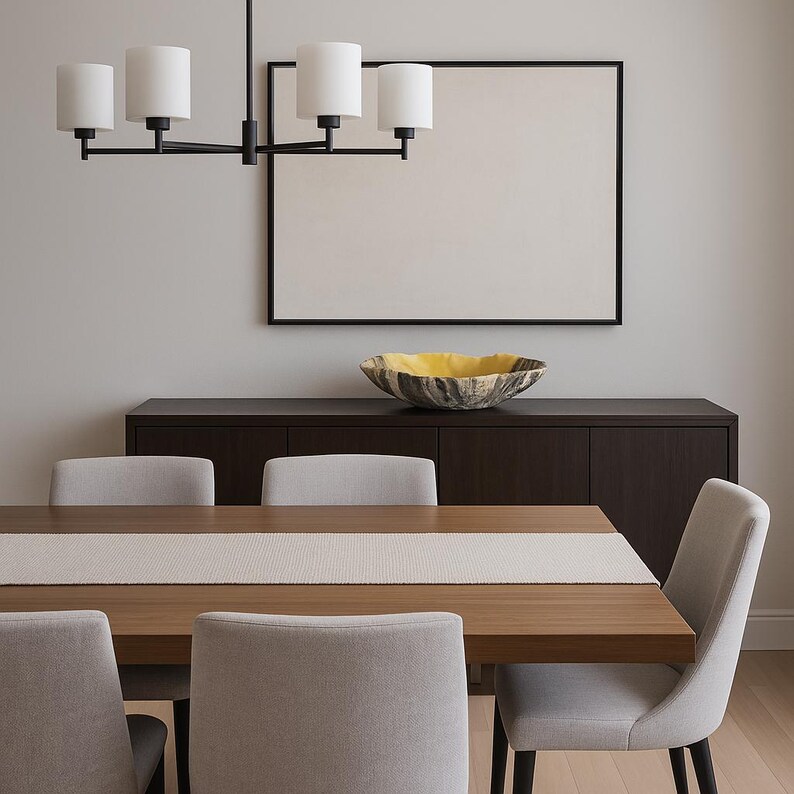 May include: A modern dining room scene featuring a wooden table with a light-colored runner, surrounded by gray upholstered chairs. A dark wood cabinet holds a decorative bowl. Above, a black framed artwork and a black chandelier with white shades.