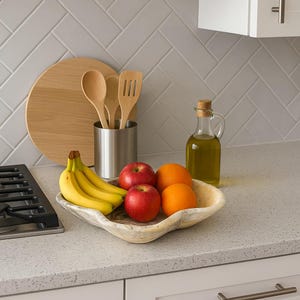 May include: A kitchen countertop scene featuring a decorative fruit bowl with bananas, red apples, and oranges. A glass bottle of olive oil, wooden utensils, and a cutting board are also present. The background includes a white tile backsplash.