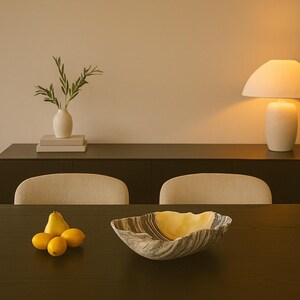 May include: A decorative bowl with a unique, wavy shape and a marbled gray and yellow pattern. The bowl sits on a dark wooden table, alongside a pear and three lemons. A white vase with greenery and a lamp are in the background.