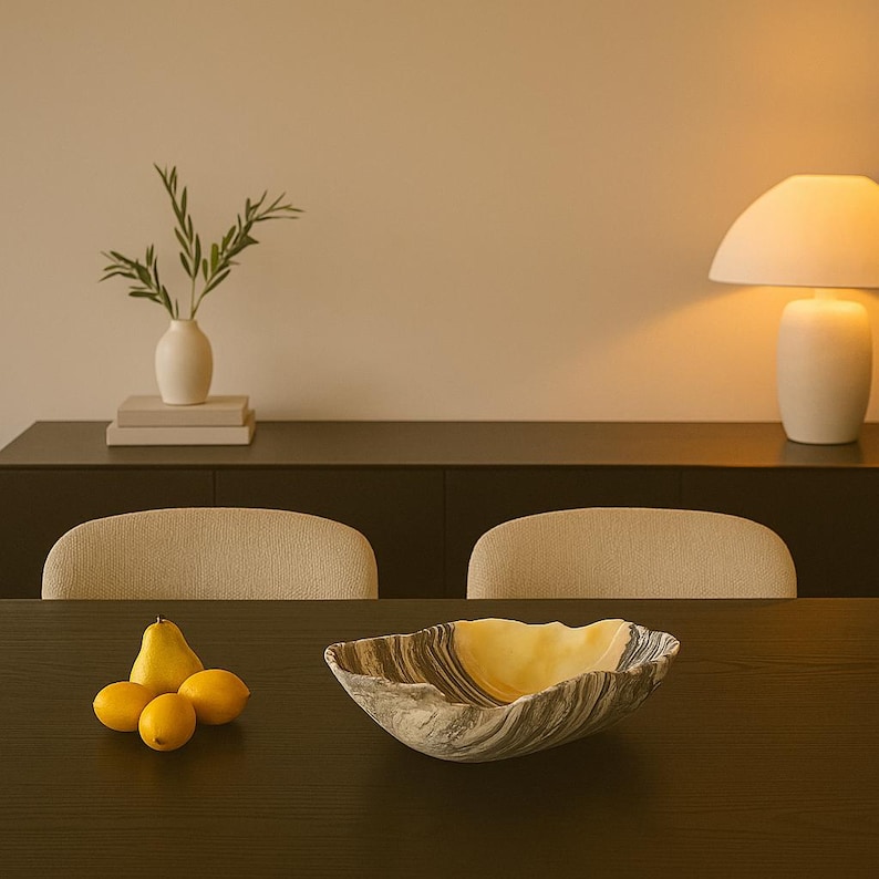 May include: A decorative bowl with a unique, wavy shape and a marbled gray and yellow pattern. The bowl sits on a dark wooden table, alongside a pear and three lemons. A white vase with greenery and a lamp are in the background.