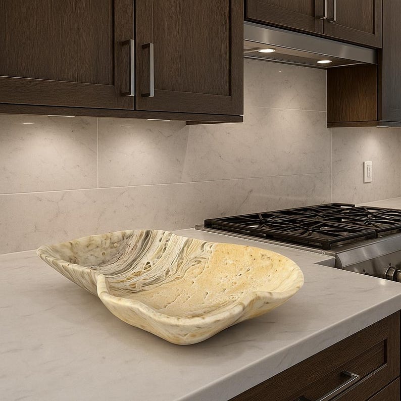 May include: A decorative, oblong bowl with a natural, swirling pattern in shades of beige, cream, and gray. The bowl is made of stone and sits on a white countertop in a kitchen setting. The bowl's shape is organic and flowing.